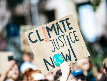 People are gathered in a crowd outside a building, and one is holding a cardboard placard saying 'Climate justice now'.