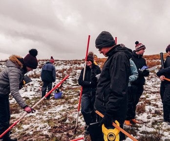 A group of 7 secondary school pupils holding ranging poles standing on a snowy hillside, they are smiling.