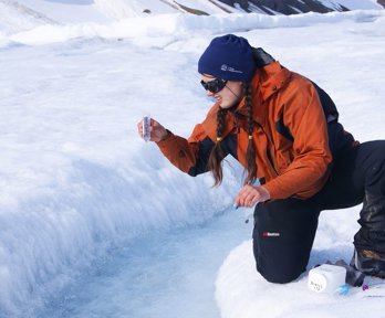 A field researcher bending down by a crack in a glacier holding a transparent tube and looking at the contents.