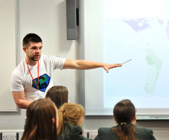Group of students watching a teacher, who's pointing at a world map on a screen.