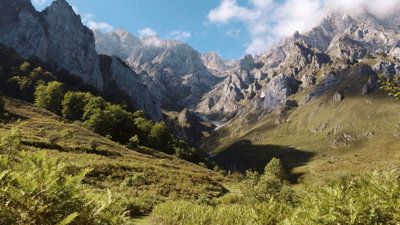 Mountain valley with steep rocky peaks, green slopes and scattered trees under a partly cloudy sky.