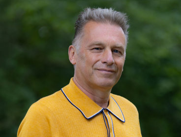 Headshot of Chris Packham wearing a bright yellow polo against a green, blurred out background.