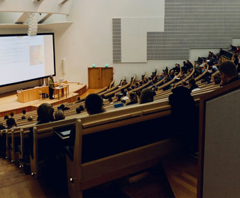 Lecture theatre with students sitting in staggered rows of seats. The rows are faced towards a stage with someone giving a presentation in front of a screen.