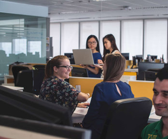Five people in office, two are talking and two are sharing a laptop