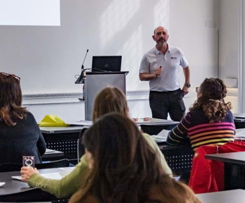 Group of people sitting at desks in a modern room listening to a presenter standing in the front.
