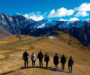A group of seven people on a brown field facing snow-covered mountains on a sunny day.