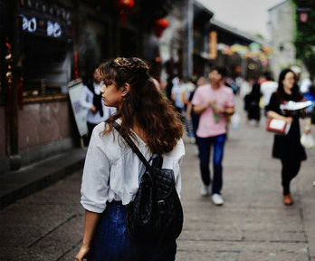 People walking on a busy street, with one person in focus as seen from behind, looking at something to their left.