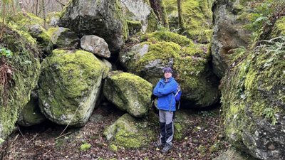 Person in blue jacket standing amongst large mossy rocks.