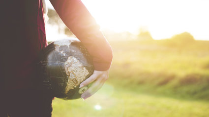 A person holding a small globe in their hand, down by their side. The sun is low in the sky and the light is bouncing off the top of the globe