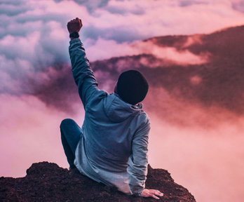 Person seen from behind sitting on a mountain cliff facing white clouds, raising a fist in the air.