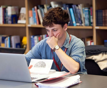 Person sat at a table in a library looking at a laptop screen with notepads and books surrounding the laptop.