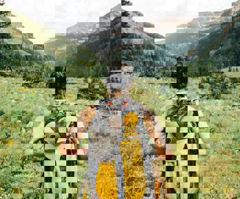 A person wearing a backpack seen from behind standing on an unpaved path surrounded by grass overlooking mountains.