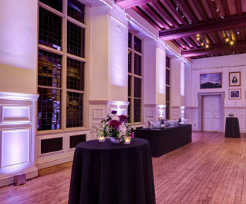 Poseur tables with floral arrangements and candles set-up for a drinks reception in a grand main hall.