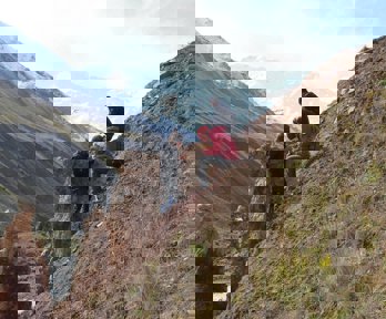 Two researchers sit on a sloping valley side while one stands, facing the others. They are all writing in notebooks.