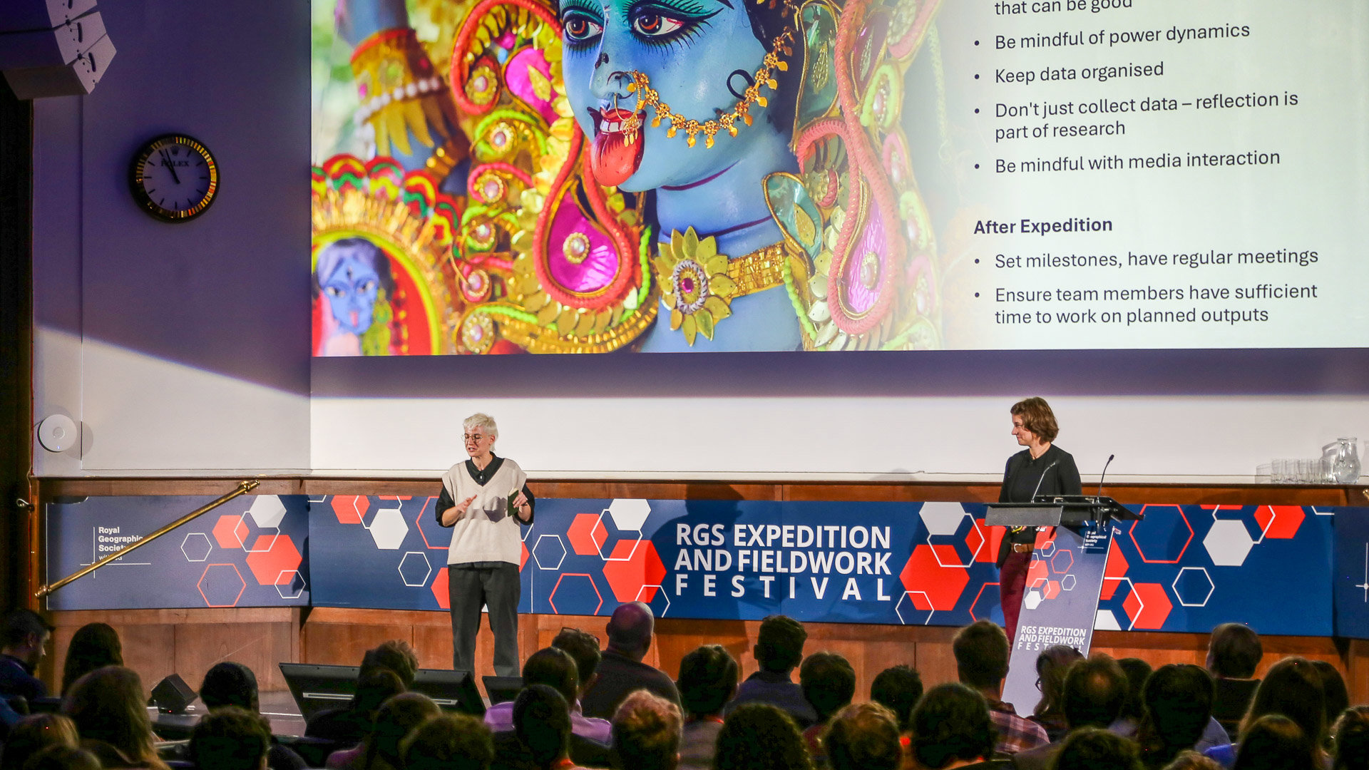 Two speakers on a stage at the RGS expedition and fieldwork festival. A slideshow with tips on how to be mindful during expeditions is projected on the wall behind them.