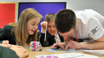 Two young children sit at their desk in a classroom looking at a small map on their desk with an adult pointing at something on the map.