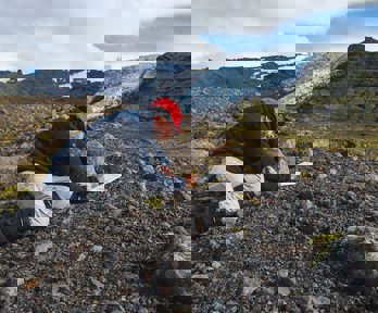Researcher kneeling in a rocky landscape writing in a notebook. There is a glacier in the background.