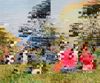 A group of young people sit on a seaside cliff, listening to a presenter in the front who's writing on a paper pad.