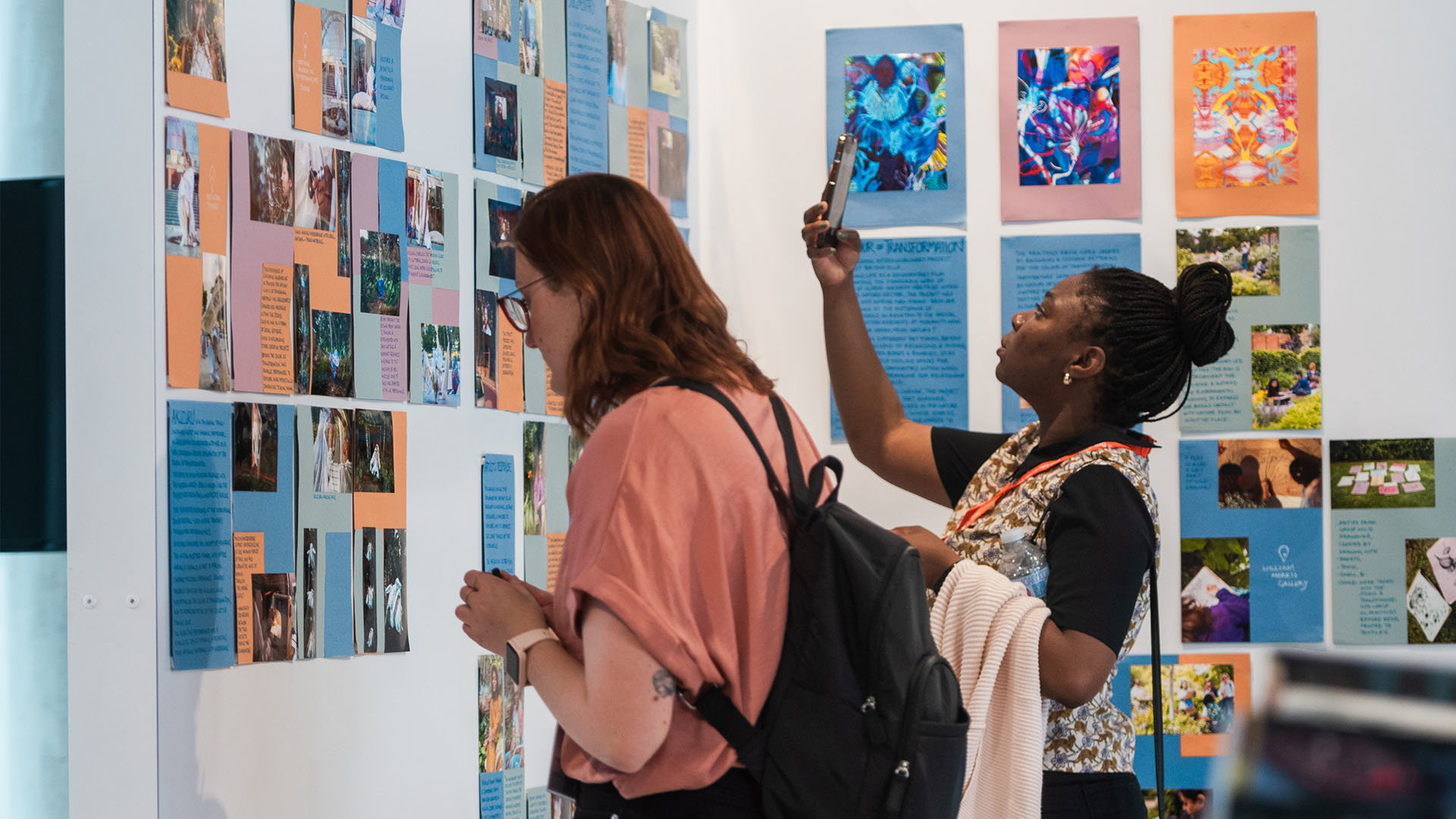 Two people stand in front of a board with posters. One is reading a caption while the other takes a photo with a smartphone.