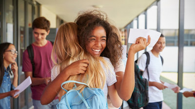 Two students hugging with exam papers in school hallway.