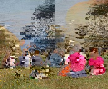 A teacher is standing at the top of a cliff, overlooking a rocky beach cove, holding a clipboard and showing a group of students something