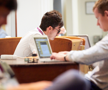 A person in the background but in focus, sits whilst looking downward. Two blurred people in the foreground also sit down, one of which is at a wooden desk typing on a laptop.