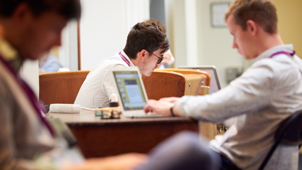 A person in the background but in focus, sits whilst looking downward. Two blurred people in the foreground also sit down, one of which is at a wooden desk typing on a laptop.