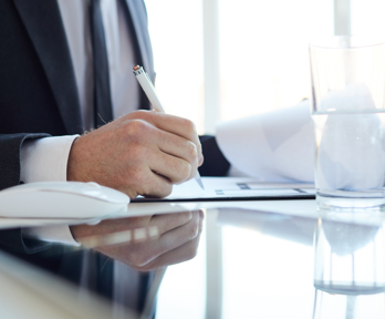 A person in a navy blue suit, white shirt and navy blue tie sits at a desk whilst using a pen to write on a spice of paper. A glass of water and white computer mouse sits next to him on the table.