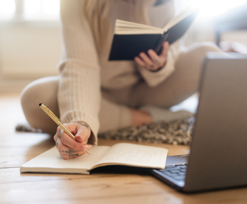 A person sits on a wooden reading a book being held in one hand and writing in a note book with their other hand. A silver laptop is laying on the ground on front of them